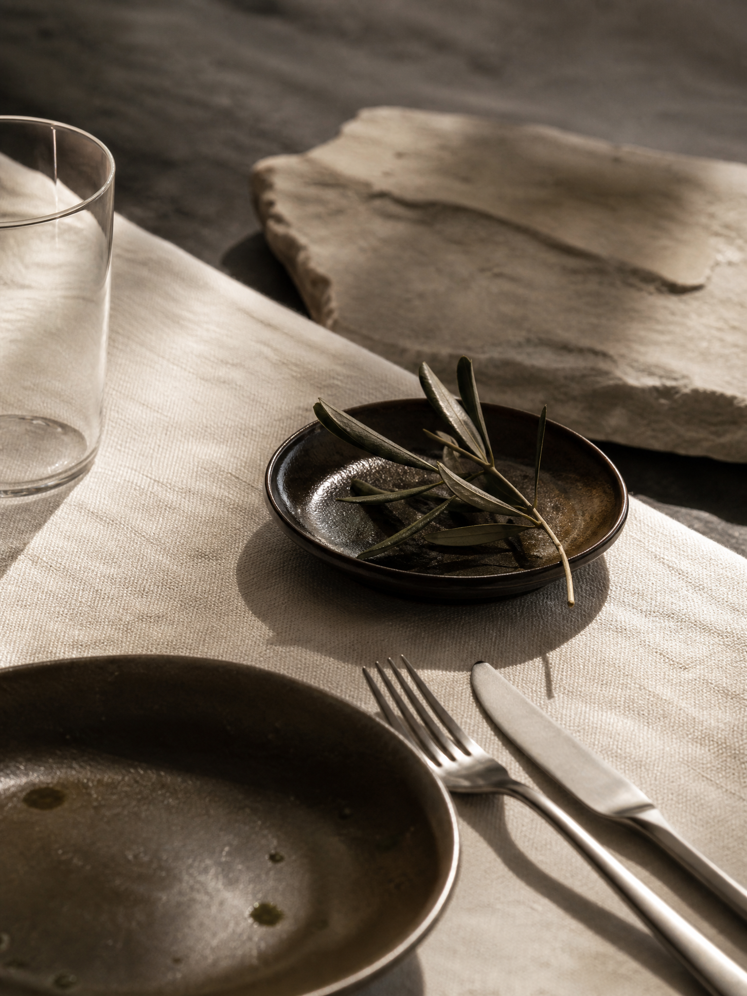 A refined still life — ivory linen, clear glass in natural light, brushed steel, and an olive branch in a small ceramic dish — evoking quiet craftsmanship