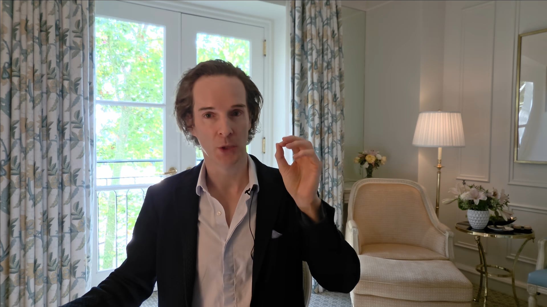 Felix Nikolas Prehn speaking in a dark jacket beside a tall window, patterned curtains and a cream armchair