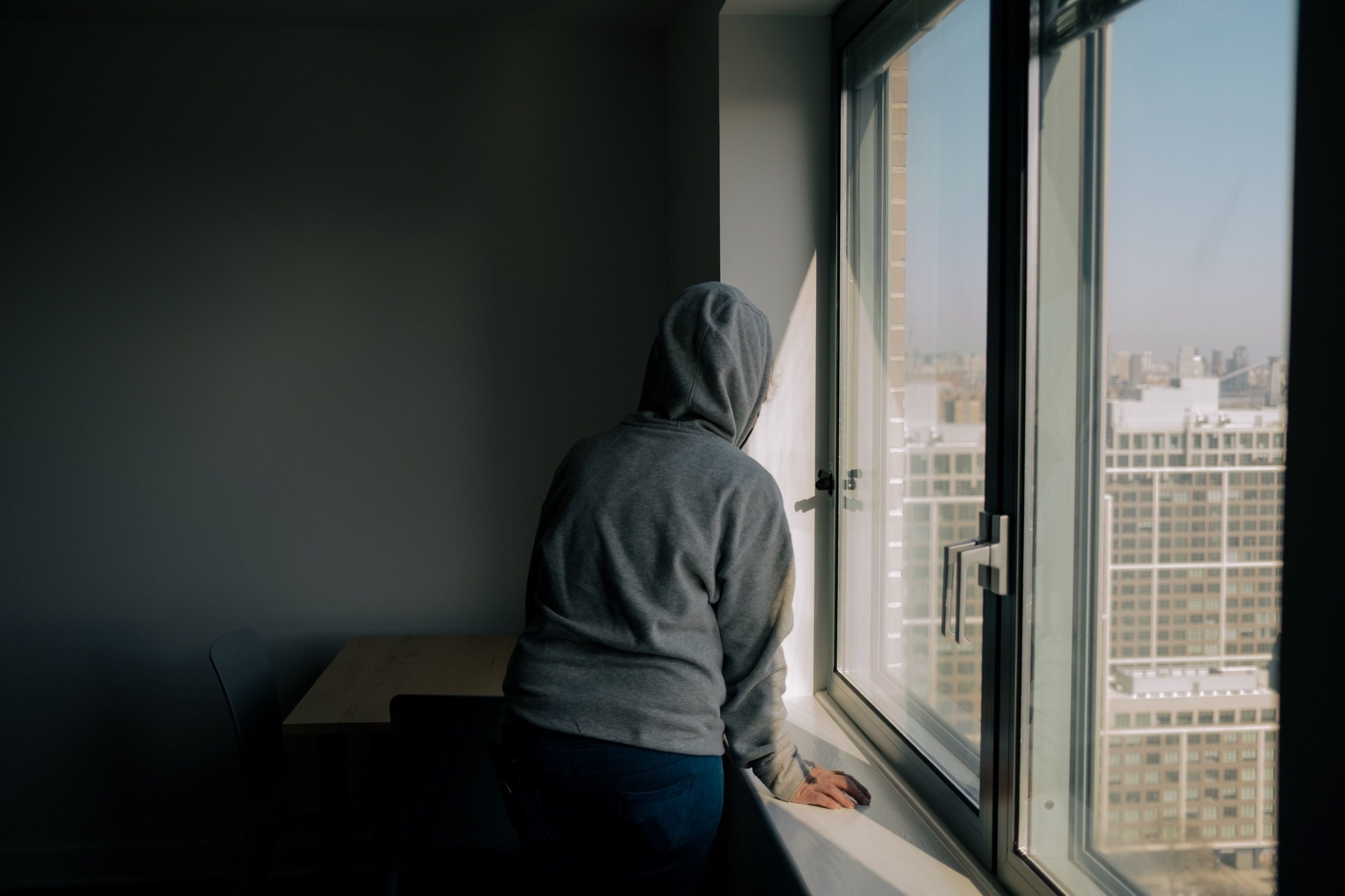 A person in a hoodie standing alone at a window in a dark room, looking out with a defeated, hopeless posture