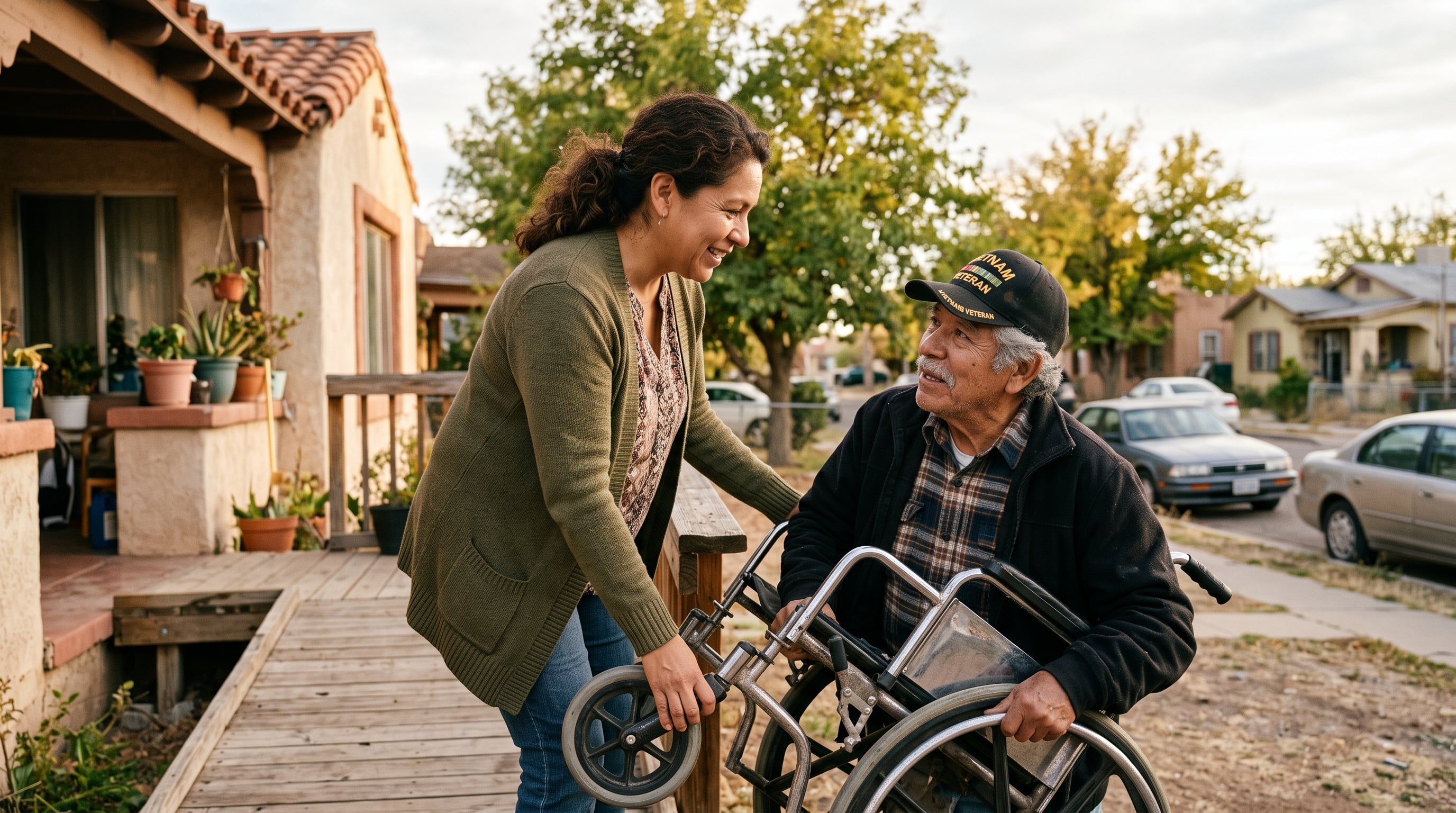 A Latina woman in her 40s helping an older Hispanic male veteran in a wheelchair up the ramp outside their home, both mid-conversation in afternoon light