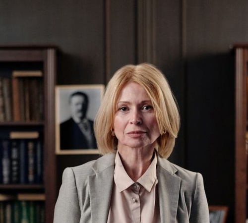 Professional white woman in her late fifties with a blonde bob wearing a gray blazer and silk blouse standing in a law office library