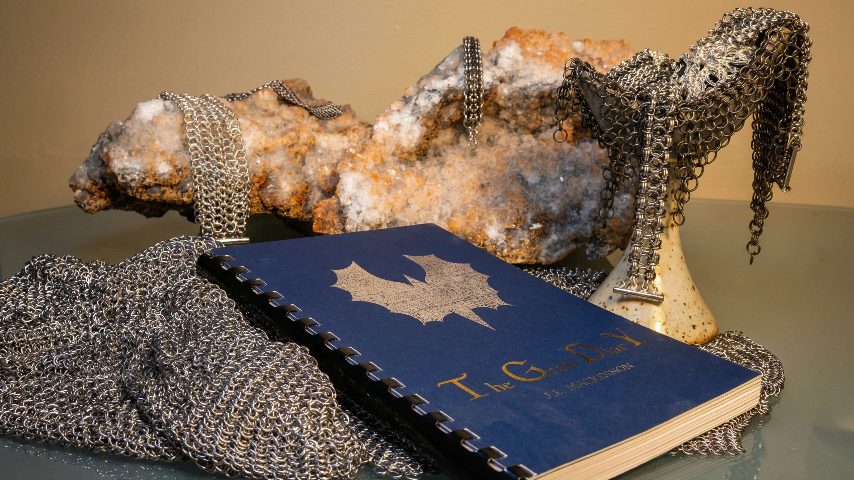 The Grail Diary Workbook — blue cover with gold maple leaf, nestled among chainmaille weaves and crystal geode