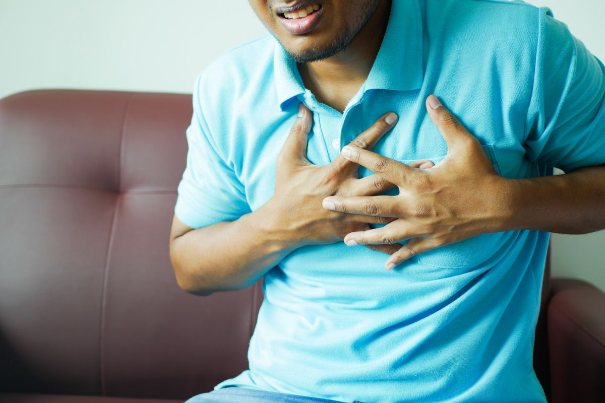 A middle-aged man sitting on a couch clutching his chest in discomfort, illustrating the cardiovascular strain caused by chronic sleep apnea