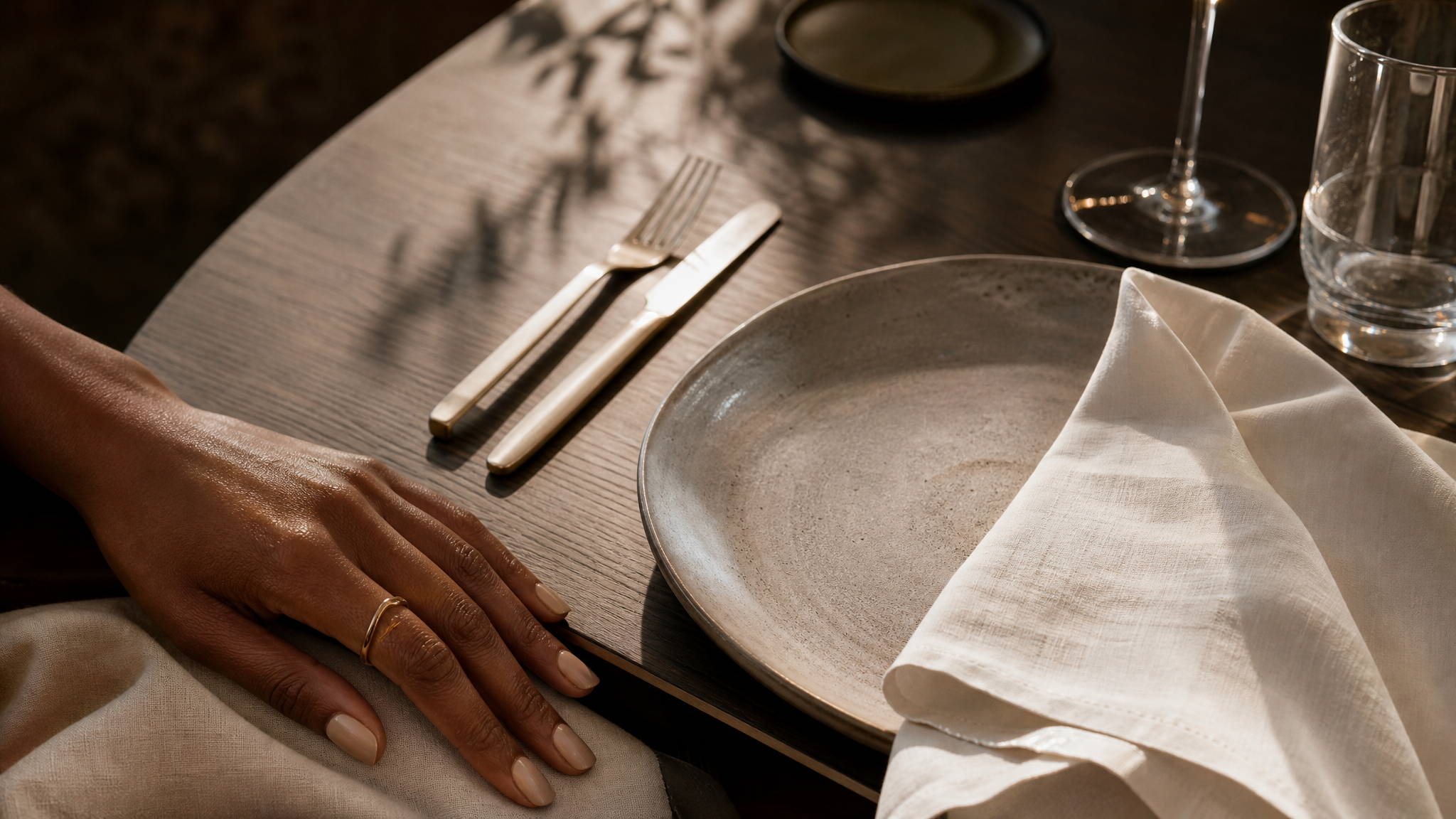 A refined table setting with crisp linen, elegant flatware, and glassware — a woman's hand resting near the table's edge, evoking arrival and a personal dining experience