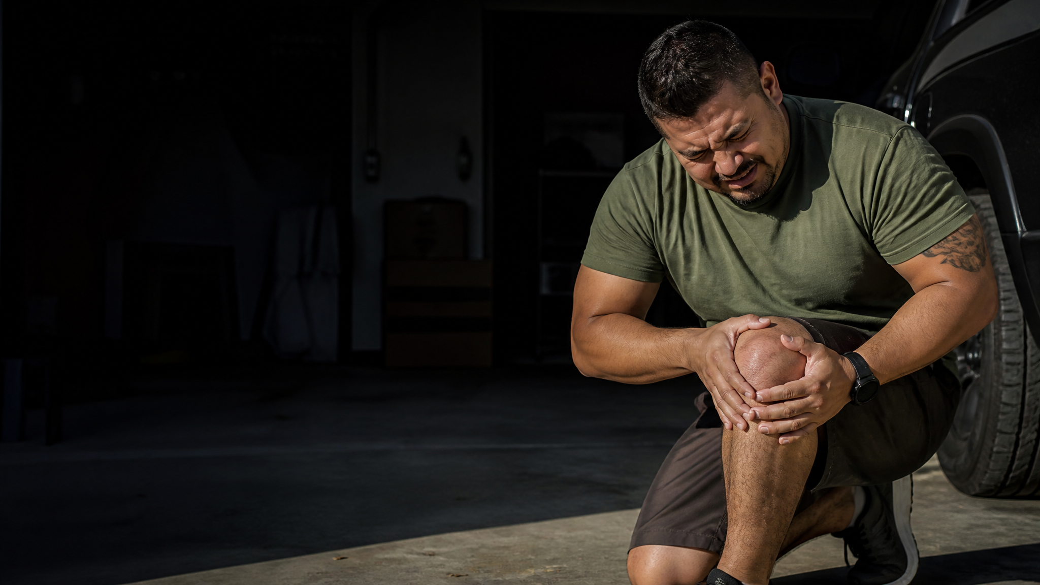 A Hispanic military veteran crouched after twisting his knee, wincing and clutching the inner side of the knee, in a garage with dramatic side light