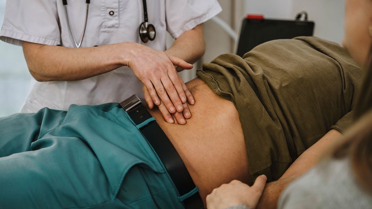 Physician in a white coat performing abdominal palpation on a patient lying on an examination table in a clinical setting