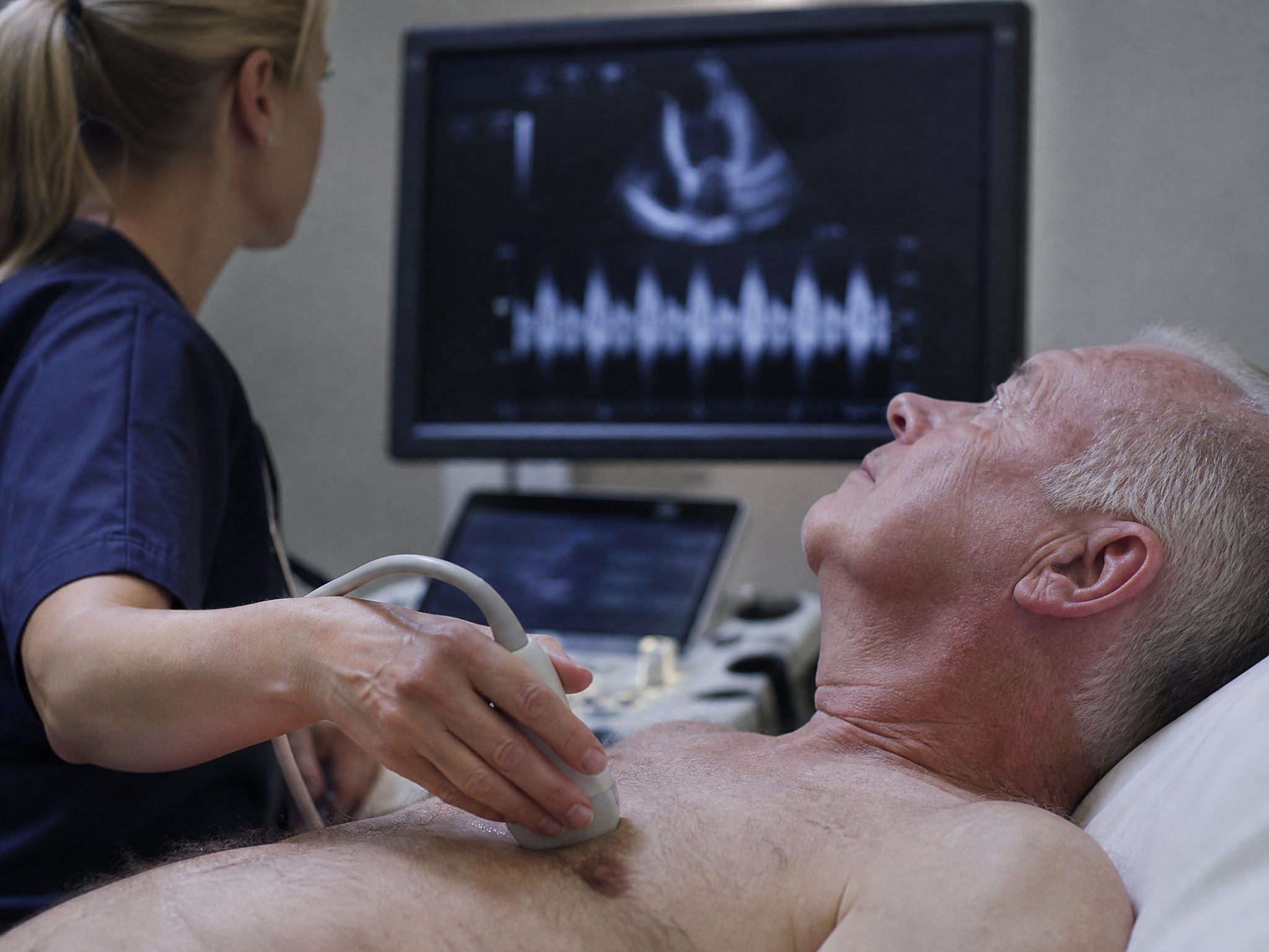 Echocardiogram procedure being performed with an ultrasound probe on a veteran chest and a monitor showing abstract ultrasound waveform patterns