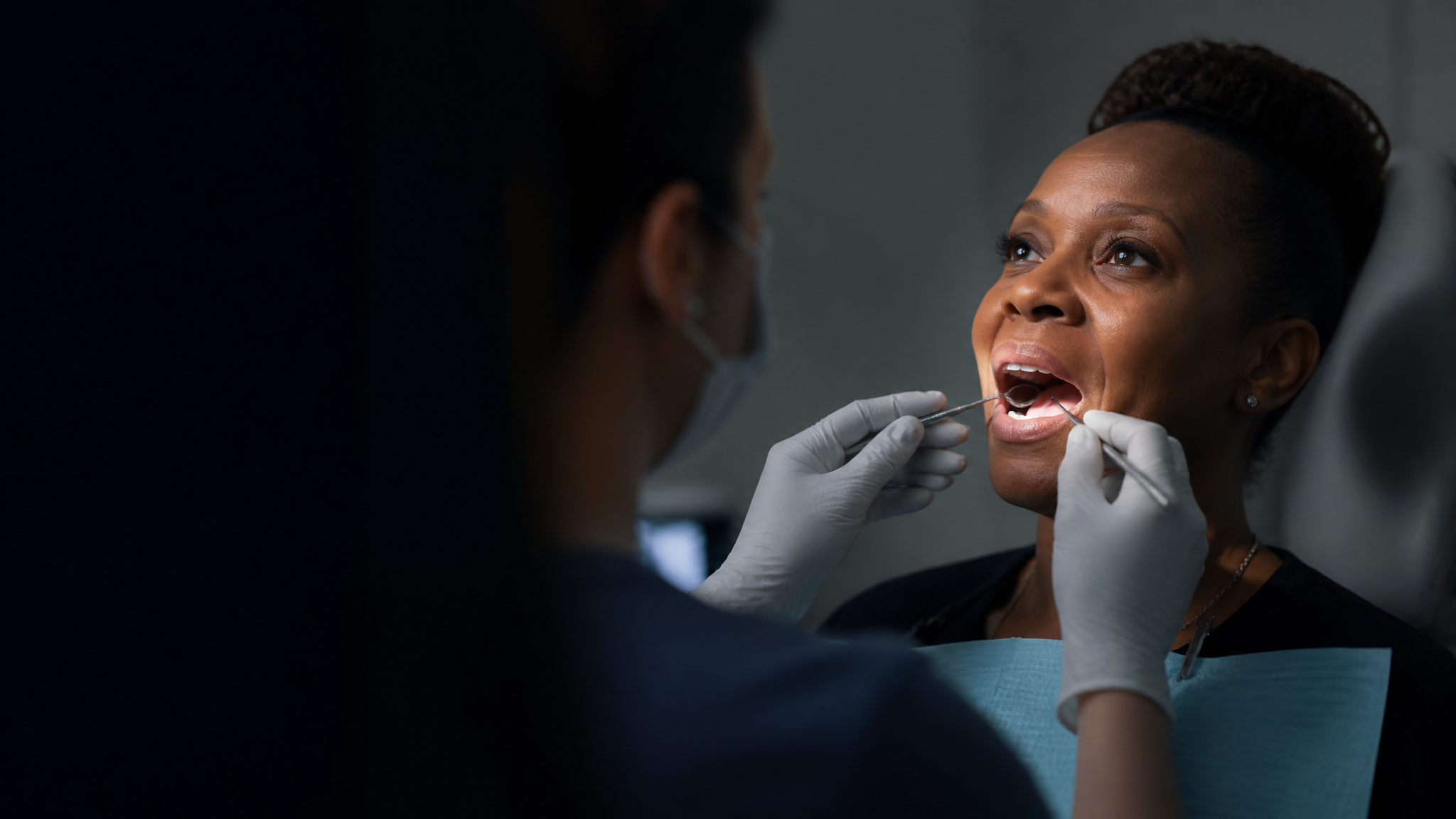 A Black female veteran in her 40s in a dental exam chair while an out-of-focus dentist examines her teeth in a dark clinical setting
