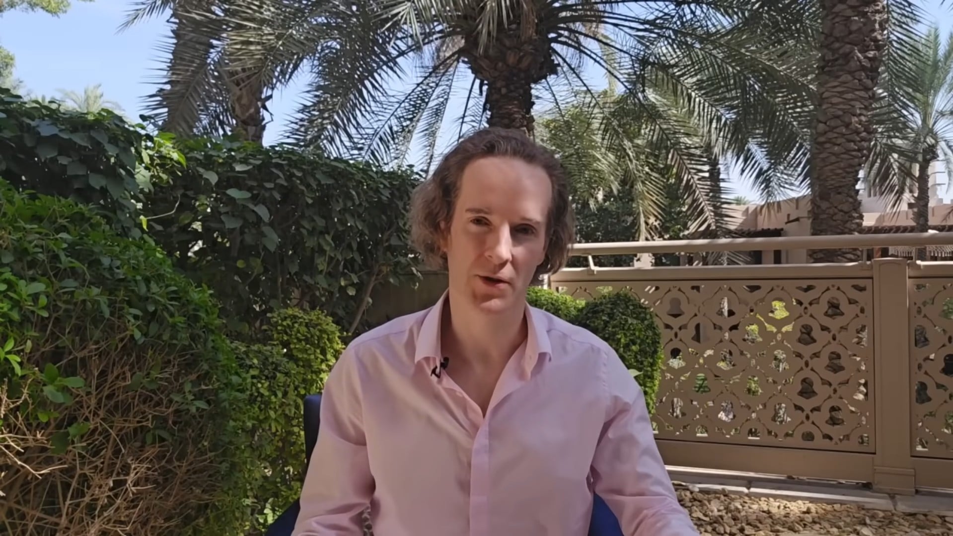 Felix Nikolas Prehn in a pink shirt in a Dubai garden, seated beside a carved railing beneath palm trees