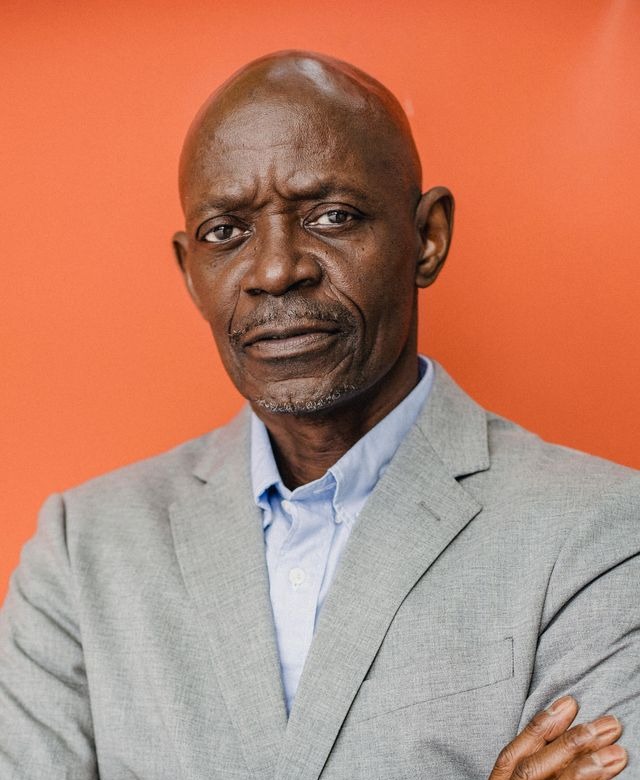 Distinguished bald Black male VA-accredited disability advocate in his mid-sixties wearing a gray suit with arms crossed in a professional portrait