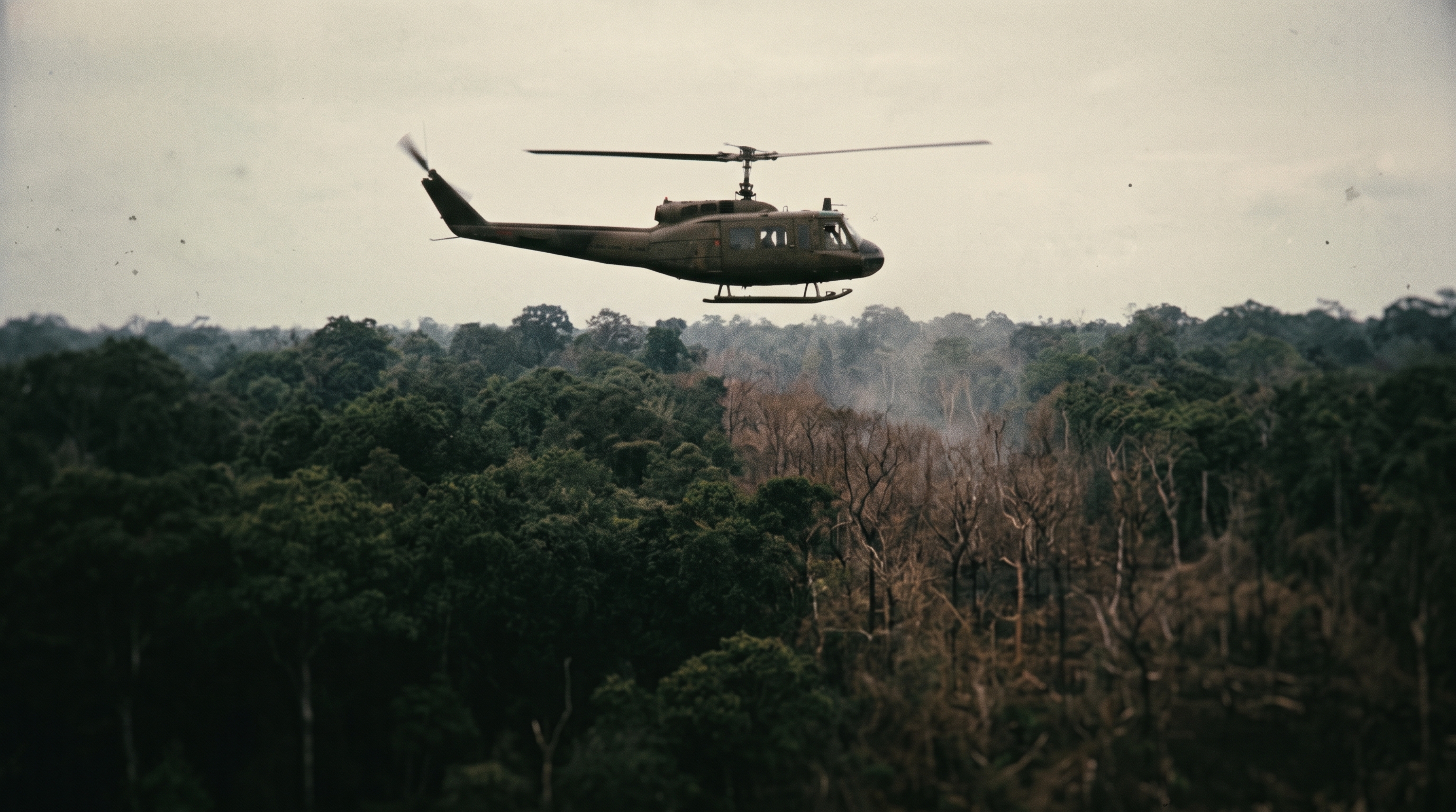 An olive-drab UH-1 Huey helicopter flying low over a defoliated Vietnam jungle treeline