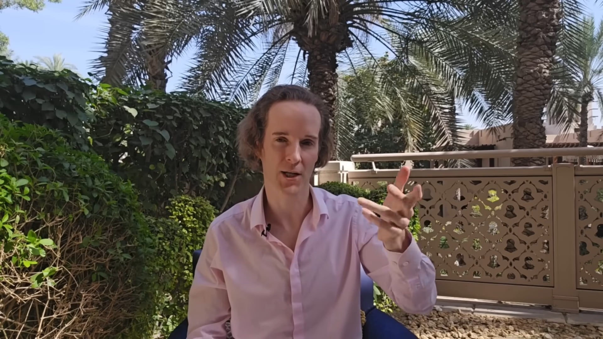 Felix Nikolas Prehn gesturing in a pink shirt in a Dubai garden, with palms and a carved railing behind him