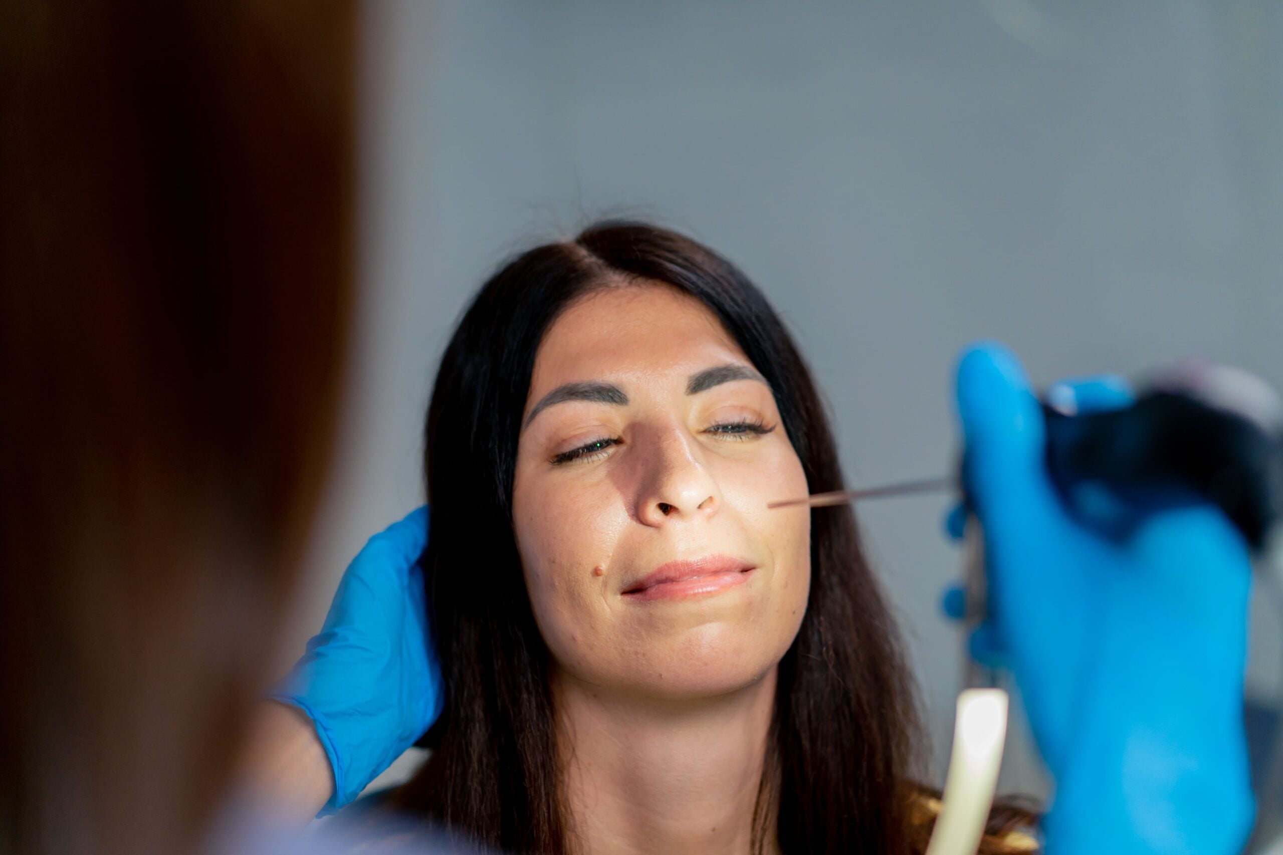 Close-up of gloved clinical hands performing a nasal endoscopy on a patient with a thin endoscope inserted into the nostril