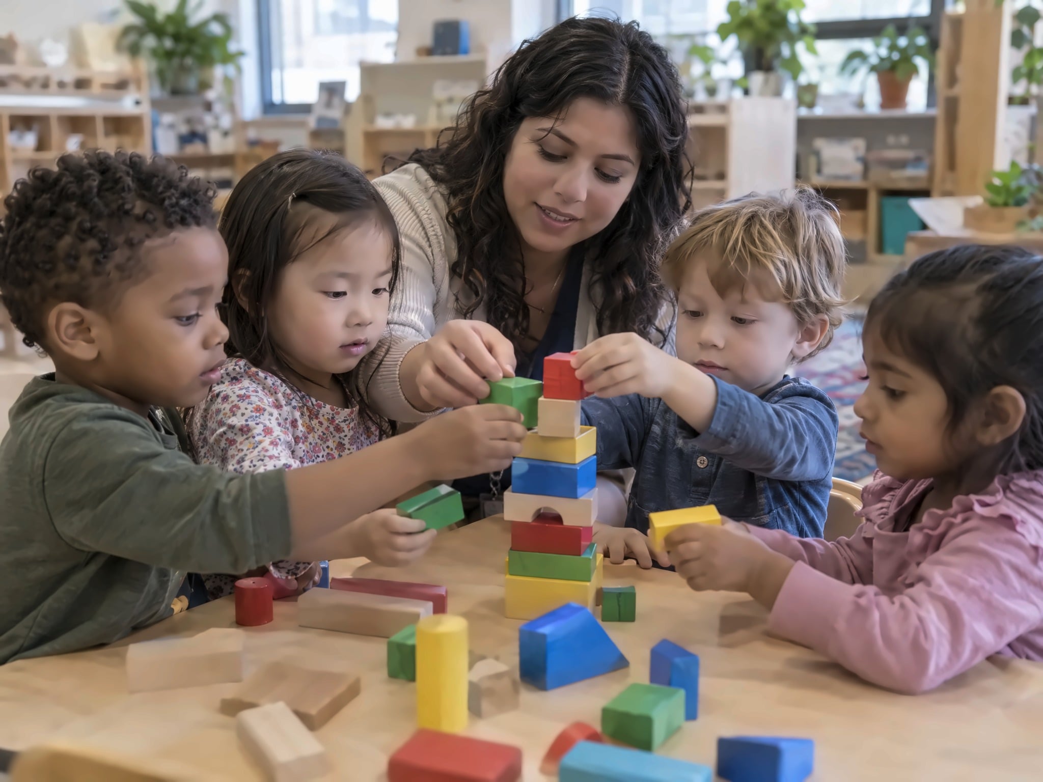 A smiling Hispanic teacher helping preschoolers build a colorful block tower