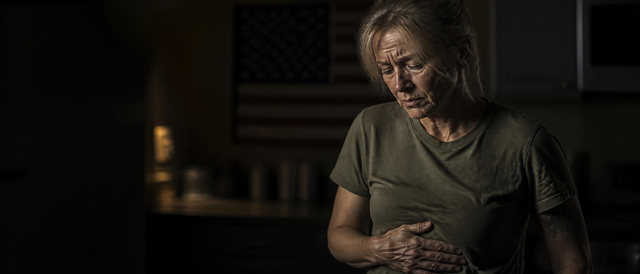 A white female military veteran in her late fifties standing in a dim kitchen at night with one hand pressed to her upper abdomen from hiatal hernia discomfort, an American flag softly out of focus behind her