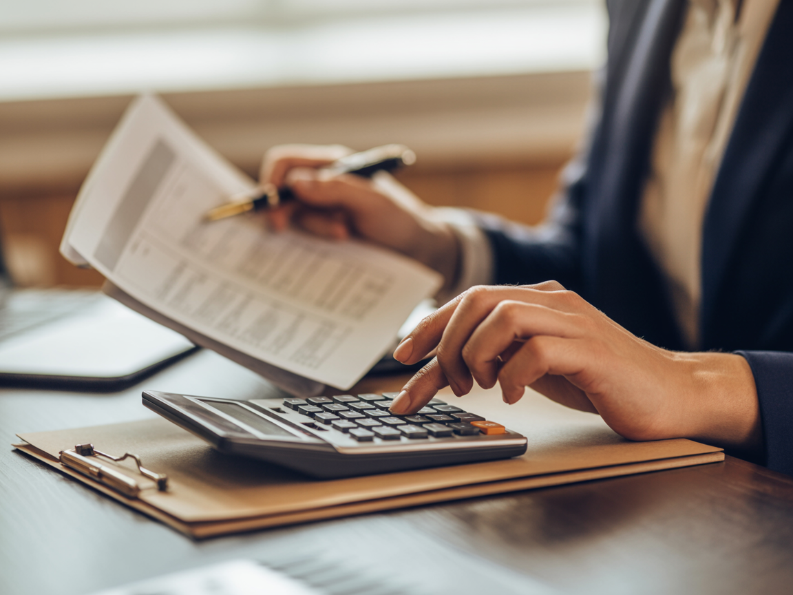 Close-up of hands using a calculator alongside spreadsheets and a pen