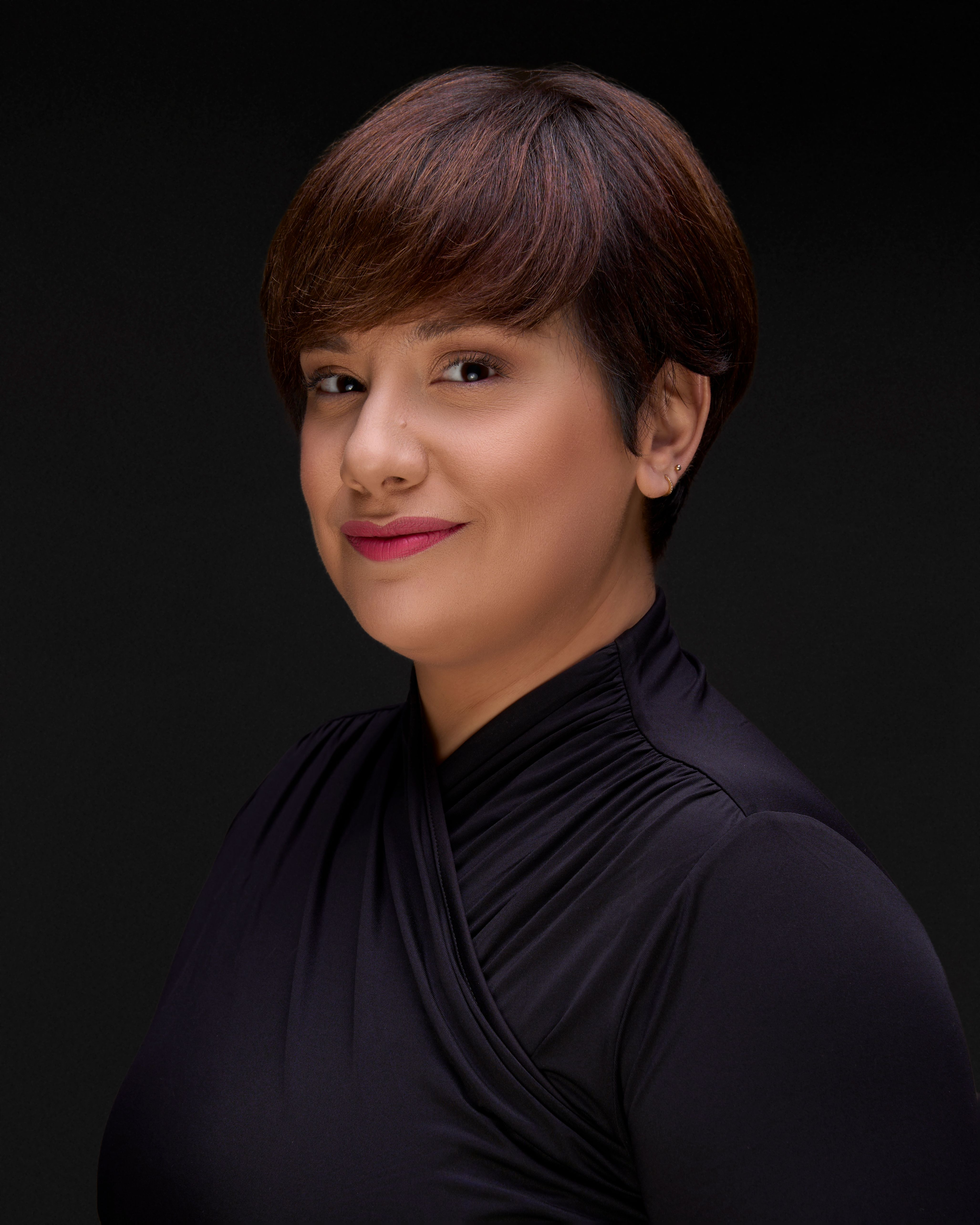 Professional South Asian woman in her mid-forties wearing a black top looking directly at the camera in a studio portrait against a dark background