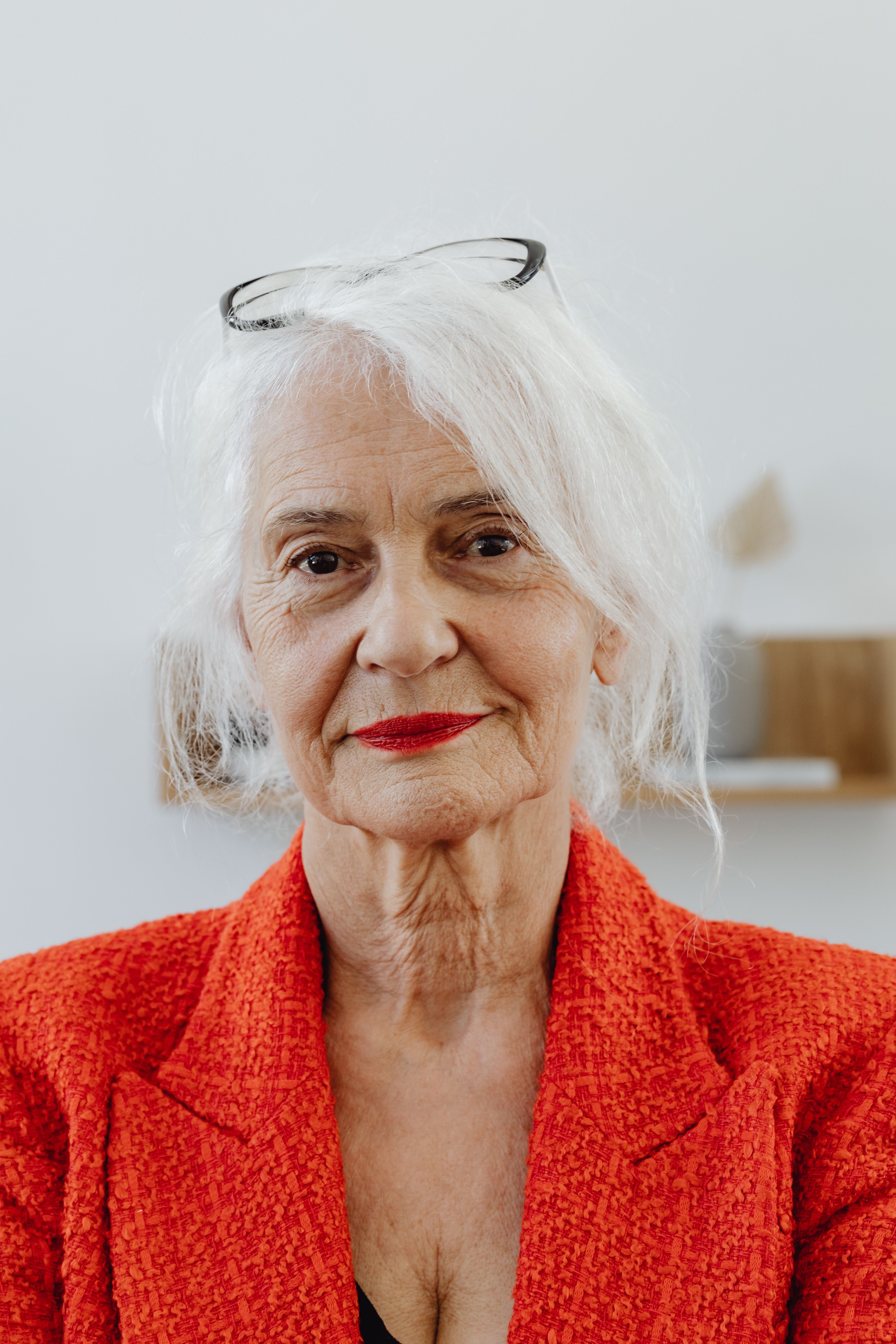A professional older white woman in a red blazer, glasses perched on her head, with a confident warm expression
