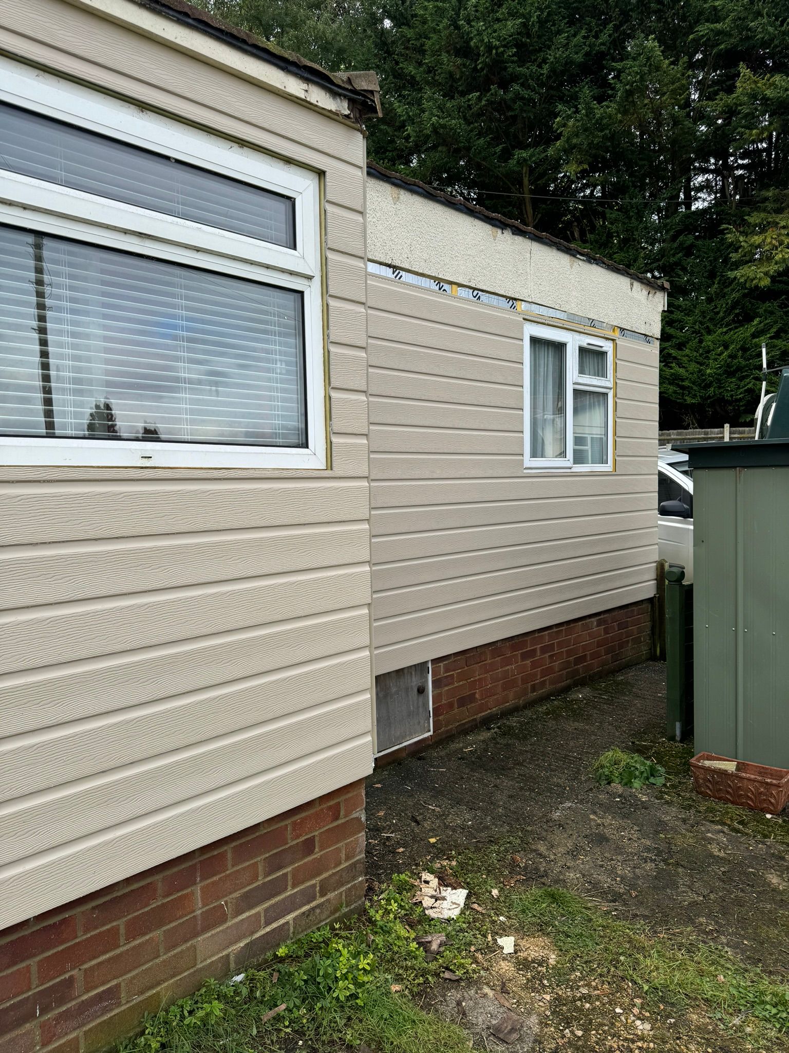 Park home corner showing completed cladding with windows and adjacent storage shed